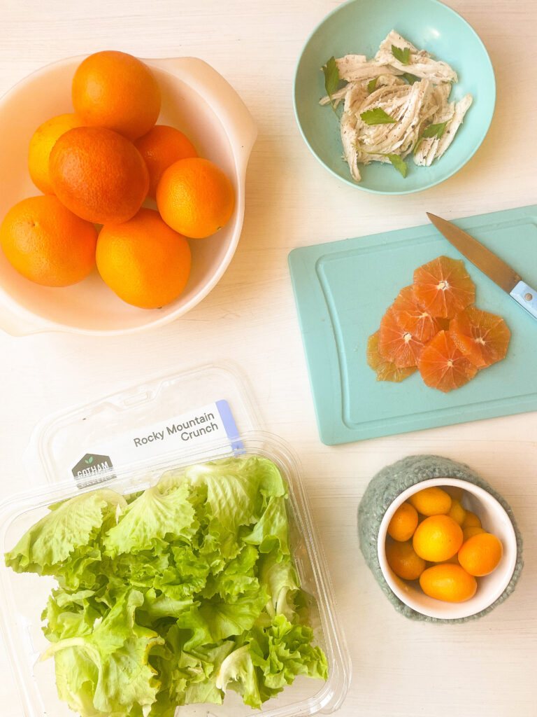 white bowl with several oranges in it, blue bowl with chicken and herbs, blue cutting board with sliced oranges sitting beside a knife, container of lettuce and small ramekin of small oranges.
