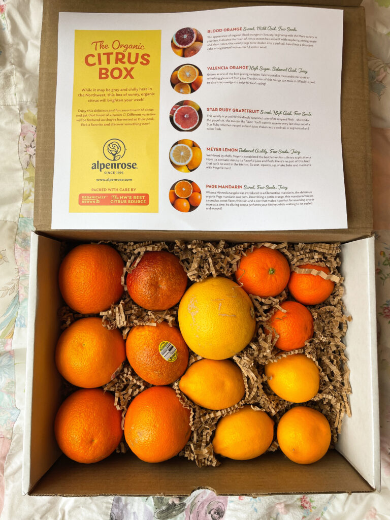Cardboard box of oranges sitting on packing paper shreds, with paper sign that lists orange types for the "Alpenrose Organic Citrus Box".