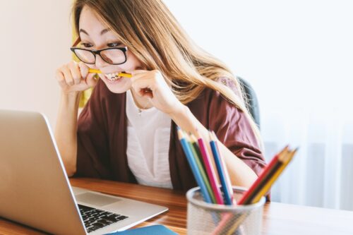 woman wearing glasses, biting on a pencil while staring at her laptop with a container of pencils on the desk next to her.