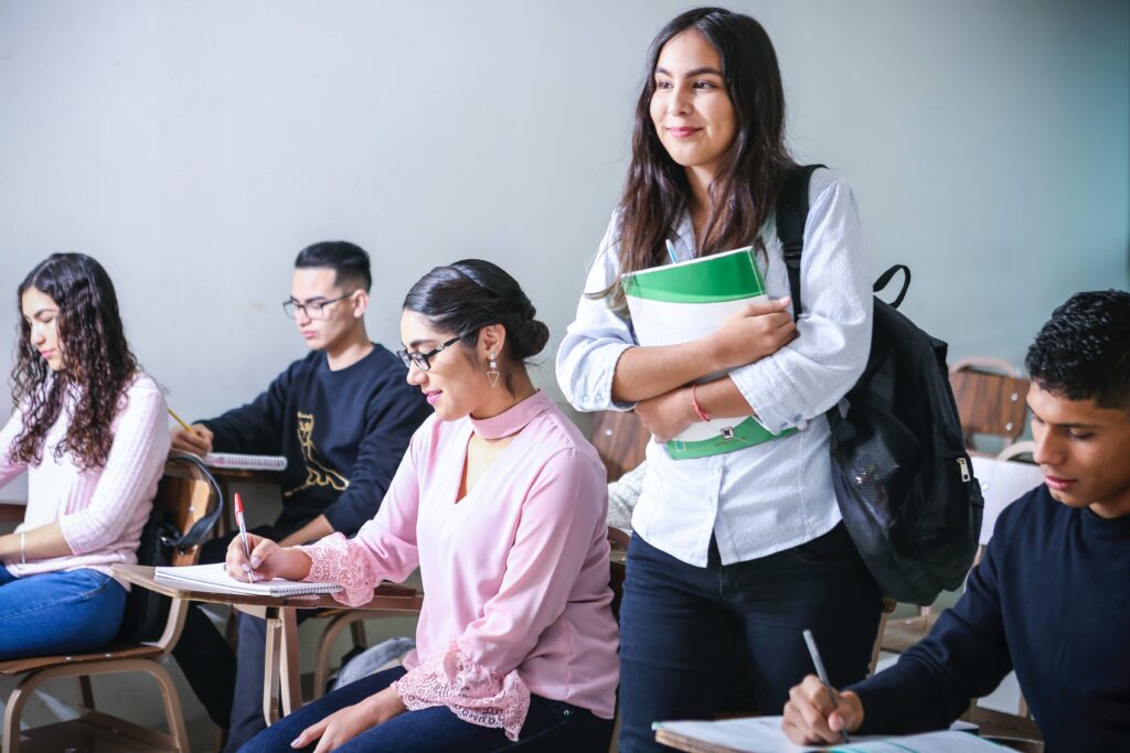 students sitting in individual desks taking notes, one student standing with backpack on and holding notebooks