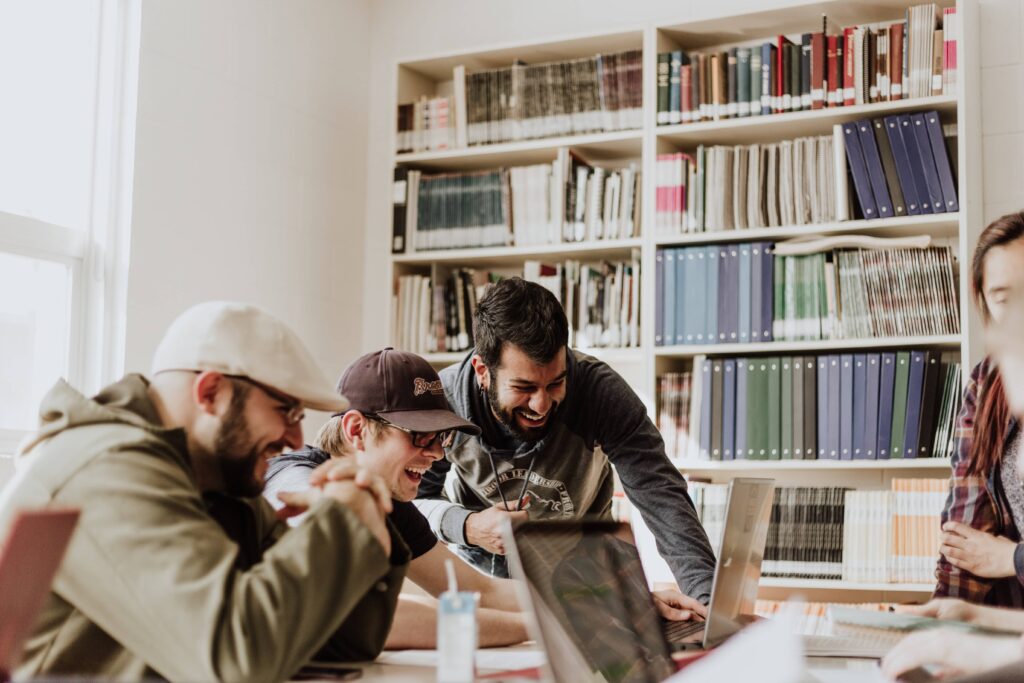 three male students laughing while looking at laptop with bookcase behind them