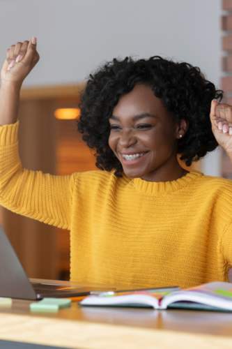 woman in yellow sweater celebrating while looking at her laptop