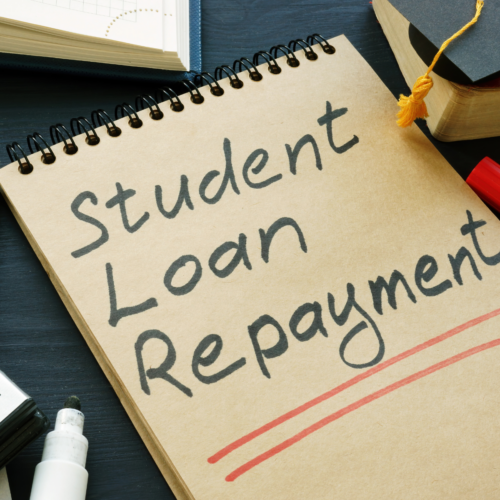 "student loan repayment" written on a tablet of paper with a book open on the table and graduation hat