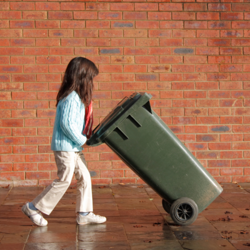 small girl walks pushing garbage bin across sidewalk doing "chores"