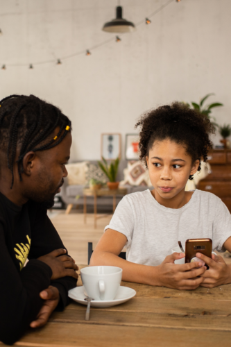 dad talking to daughter about finances at the kitchen table while dad drinks coffee and child holds cell phone