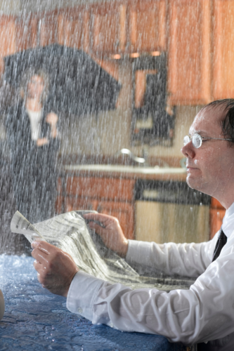 man reading paper in "rain" in his kitchen. Woman behind him with an umbrella